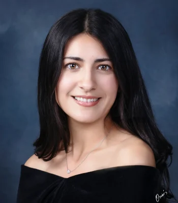 Senior portrait of a smiling young woman with long dark hair and a delicate pendant necklace, wearing an off-the-shoulder black drape against a blue backdrop.