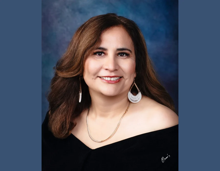 Portrait of a smiling woman with auburn-brown hair, large white crochet teardrop earrings and a gold chain, wearing an off-the-shoulder black drape.
