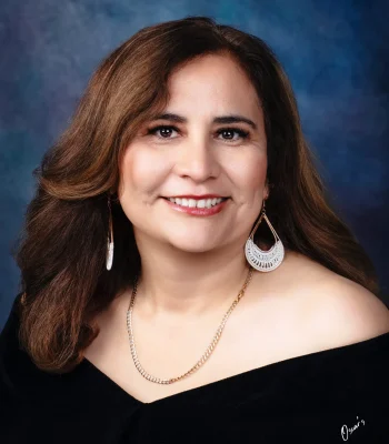 Portrait of a smiling woman with auburn-brown hair, large white crochet teardrop earrings and a gold chain, wearing an off-the-shoulder black drape.