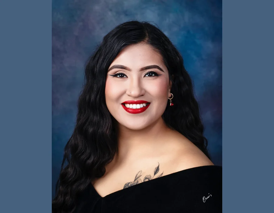 Senior portrait of a smiling young woman with wavy black hair, bold red lipstick and a floral shoulder tattoo, wearing an off-the-shoulder black drape against a blue backdrop.