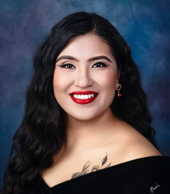Senior portrait of a smiling young woman with wavy black hair, bold red lipstick and a floral shoulder tattoo, wearing an off-the-shoulder black drape against a blue backdrop.