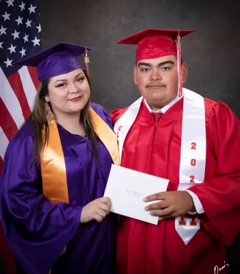 Two graduates standing together, a young woman in a purple cap and gown with a gold stole and a young man in a red cap and gown with a white class stole, holding a Hueneme High School diploma in front of an American flag.