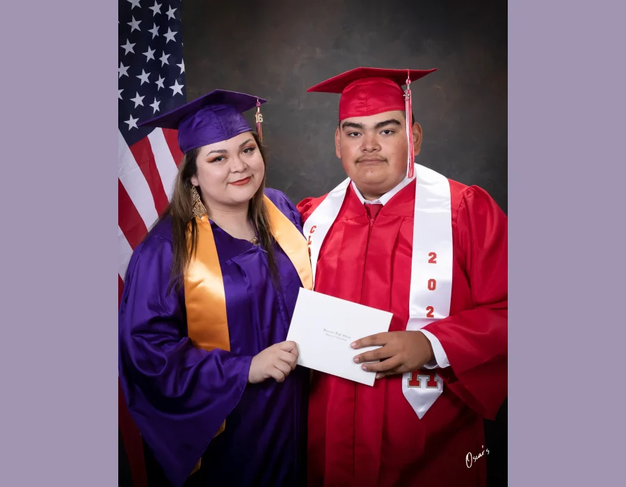 Two graduates standing together, a young woman in a purple cap and gown with a gold stole and a young man in a red cap and gown with a white class stole, holding a Hueneme High School diploma in front of an American flag.