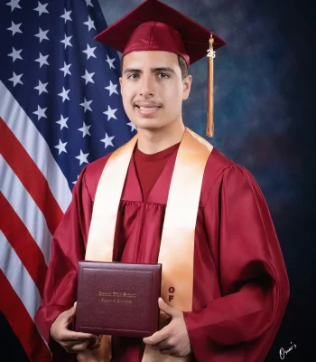 Young man in a maroon cap and gown with a peach stole and a "26" tassel charm, holding an Oxnard High School diploma in front of an American flag at Oscar's Photography and Video.