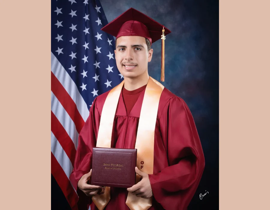 Young man in a maroon cap and gown with a peach stole and a "26" tassel charm, holding an Oxnard High School diploma in front of an American flag at Oscar's Photography and Video.