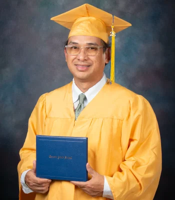 Adult graduate in glasses, a gold cap and gown and a tie, smiling and holding a blue Oxnard Adult School diploma cover.