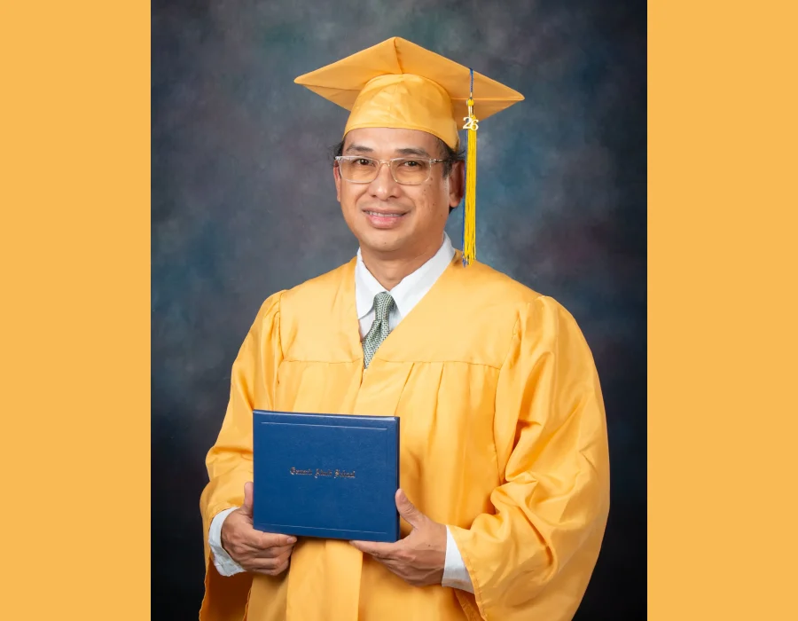 Adult graduate in glasses, a gold cap and gown and a tie, smiling and holding a blue Oxnard Adult School diploma cover.