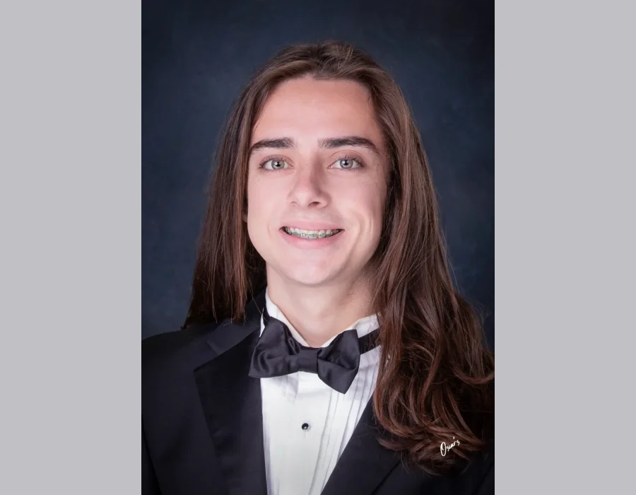 Senior portrait of a smiling young man with long brown hair and braces, wearing a black tuxedo and bow tie against a dark blue backdrop.