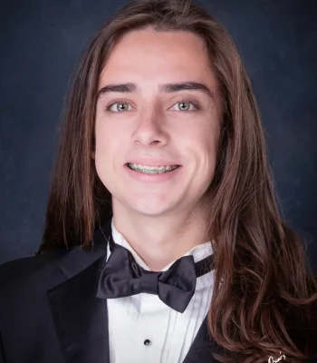 Senior portrait of a smiling young man with long brown hair and braces, wearing a black tuxedo and bow tie against a dark blue backdrop.
