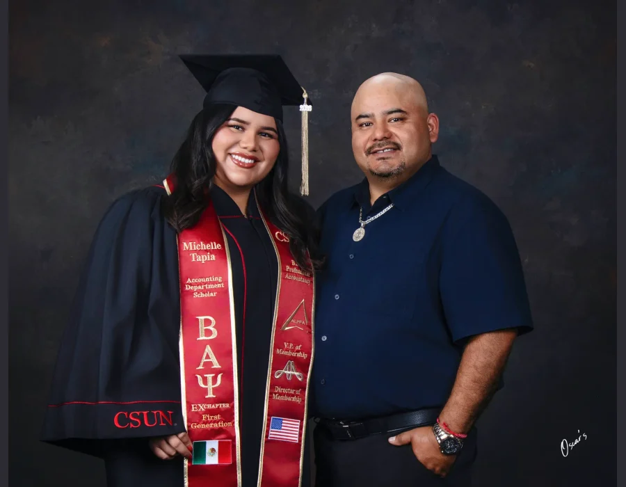 Smiling CSUN accounting graduate in her cap, gown and embroidered red stole, posing beside her father in a navy button-up shirt.