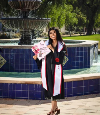 Joyful graduate in a black gown and white stole holding up a decorated cap reading "Gracias Papi y Mami por su apoyo, amor y sacrificios. Lo logramos," beside a tiled campus fountain.
