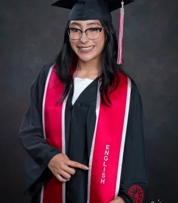 Smiling graduate in glasses, a black cap and gown, and a red stole embroidered "English," pointing to her major in a studio portrait.
