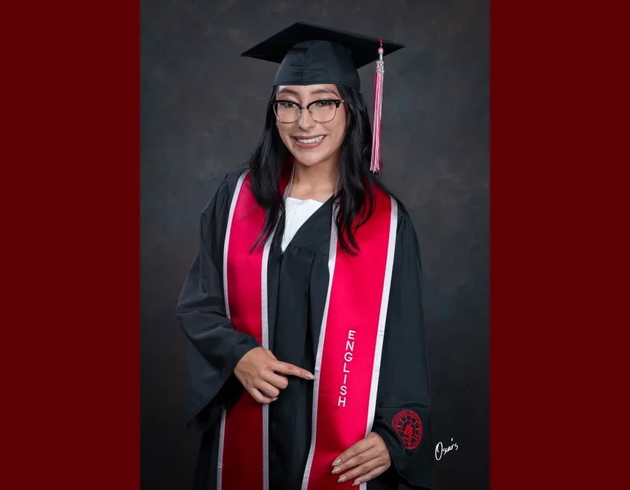 Smiling graduate in glasses, a black cap and gown, and a red stole embroidered "English," pointing to her major in a studio portrait.