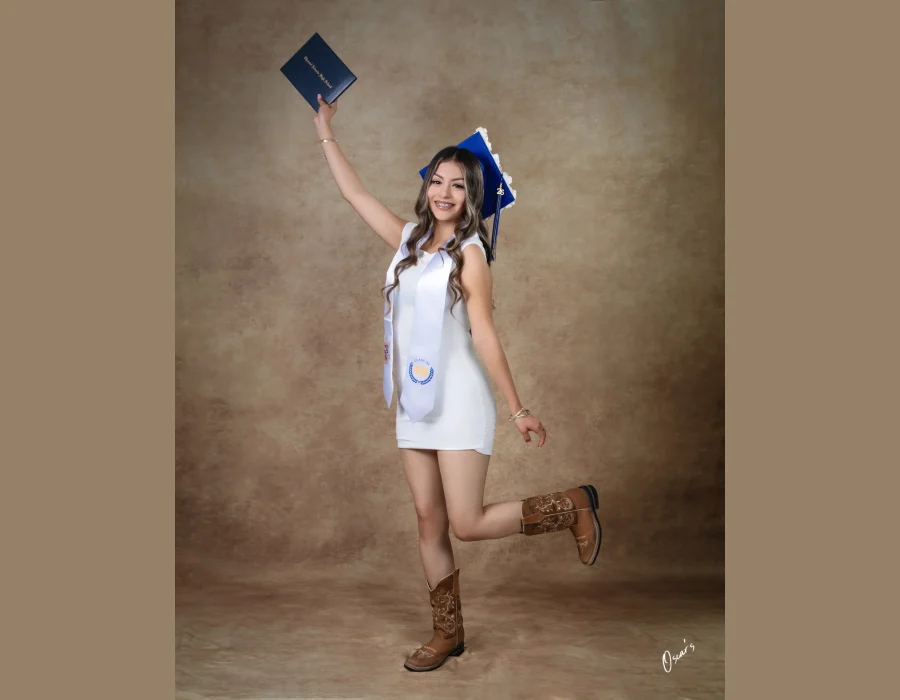 Graduation portrait of a girl in a white dress and cowboy boots jumping with her diploma raised, Class of 2026 sash
