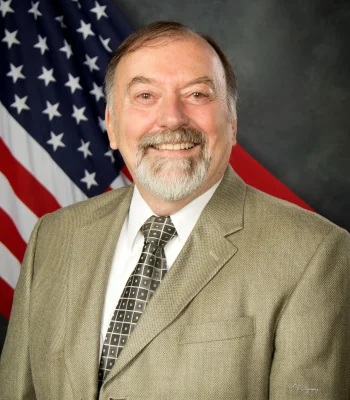 Professional corporate headshot of an older man in a tan blazer and patterned tie, studio portrait with American flag