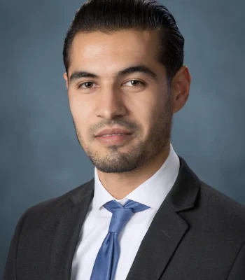 Professional corporate headshot of a young man in a dark suit and blue tie, studio portrait