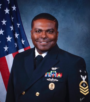 Portrait of a Navy service member in dress uniform with medals and rank insignia, studio backdrop with American flag