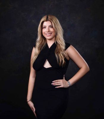 Professional portrait of a woman in a black dress against a dark studio backdrop, hand on hip