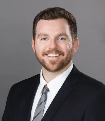Professional corporate headshot of a man in a dark suit and striped tie, studio gray background