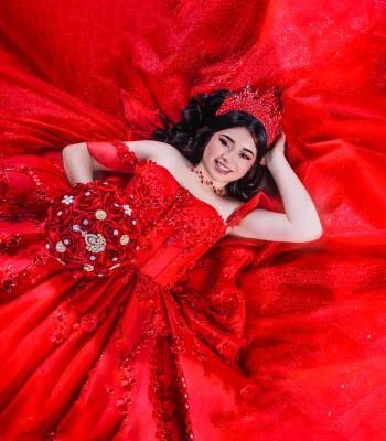 Quinceañera overhead portrait lying on a red satin gown with a jeweled crown, smiling while holding a red rose bouquet