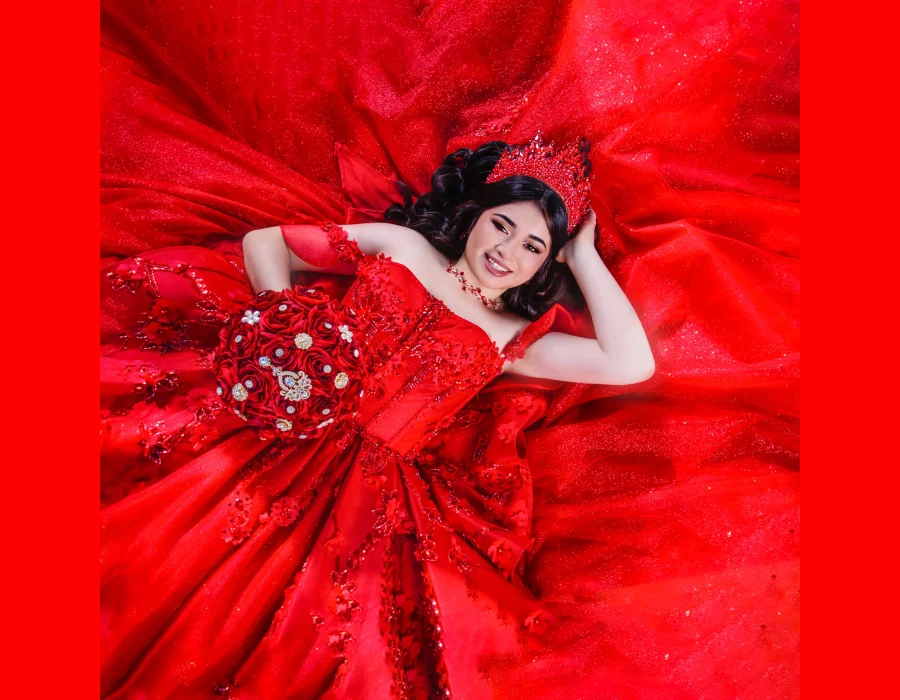 Quinceañera overhead portrait lying on a red satin gown with a jeweled crown, smiling while holding a red rose bouquet