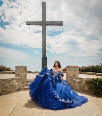 Quinceañera portrait in a royal blue sequined gown with silver embroidery, standing before a large wooden cross overlook