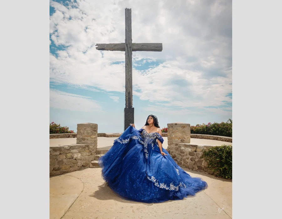 Quinceañera portrait in a royal blue sequined gown with silver embroidery, standing before a large wooden cross overlook