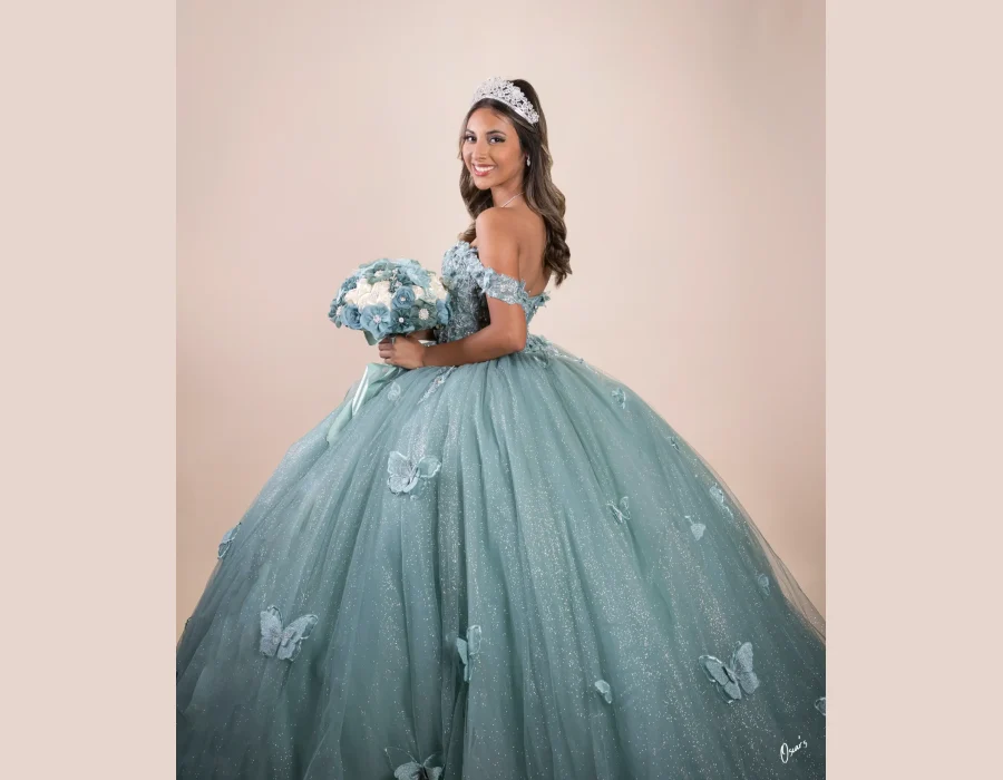 Quinceañera portrait in a sage green beaded gown, smiling over her shoulder against a soft pink studio backdrop