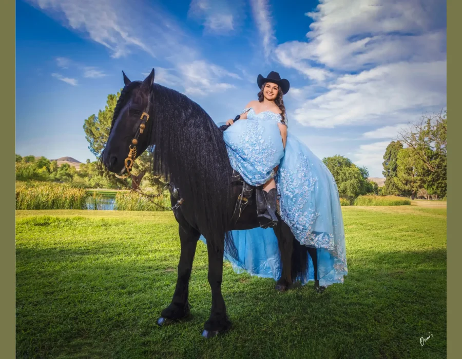 Quinceañera portrait riding a black horse sidesaddle in a light blue beaded gown and black cowboy hat