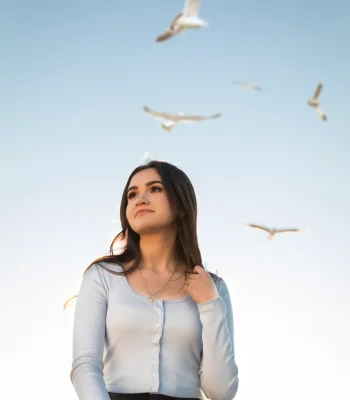 Quinceañera senior-style portrait in a light blue top, looking up at seagulls flying overhead near the beach