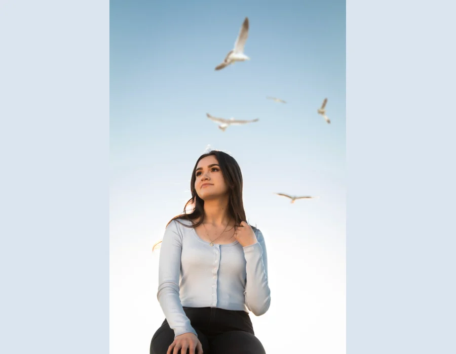 Quinceañera senior-style portrait in a light blue top, looking up at seagulls flying overhead near the beach