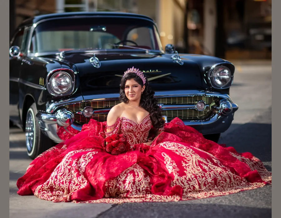 Quinceañera portrait in a red beaded gown with a long ruffled train, posing beside a vintage black classic car