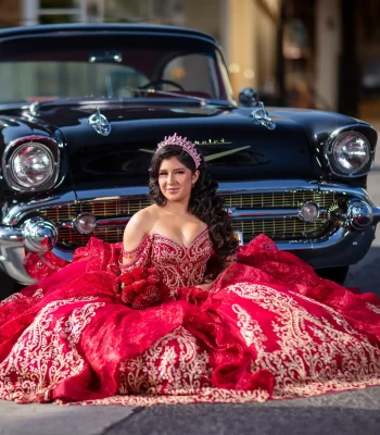 Quinceañera portrait in a red beaded gown with a long ruffled train, posing beside a vintage black classic car