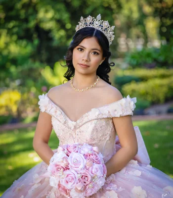 Quinceañera close-up portrait in a blush off-the-shoulder gown with floral trim and a gold tiara, holding a pink rose bouquet