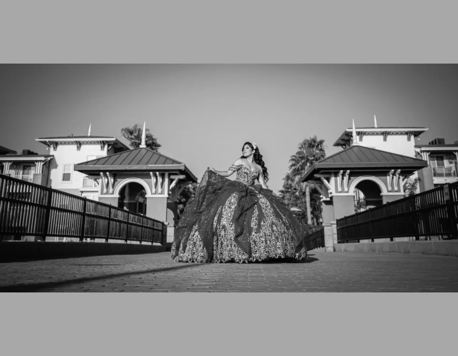 Quinceañera black-and-white portrait in a dark embellished ball gown, walking across a bridge between two gazebos