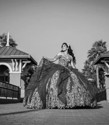 Quinceañera black-and-white portrait in a dark embellished ball gown, walking across a bridge between two gazebos