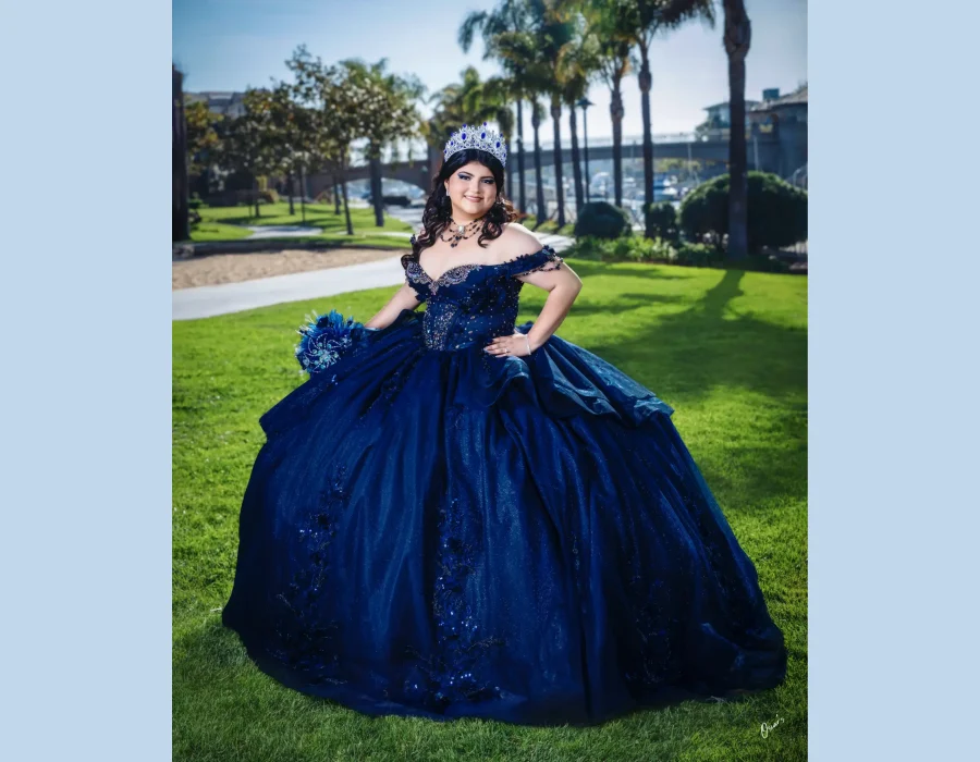 Quinceañera full-length portrait in a navy blue sequined gown with a sapphire tiara, posing on a lawn near palm trees and a marina