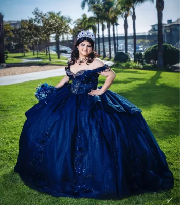 Quinceañera full-length portrait in a navy blue sequined gown with a sapphire tiara, posing on a lawn near palm trees and a marina
