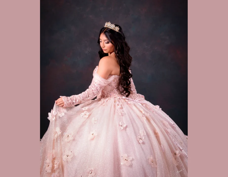 Quinceañera portrait in a blush pink off-the-shoulder ball gown with floral appliqués, wearing a gold tiara, looking over her shoulder against a dark studio backdrop