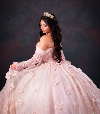 Quinceañera portrait in a blush pink off-the-shoulder ball gown with floral appliqués, wearing a gold tiara, looking over her shoulder against a dark studio backdrop