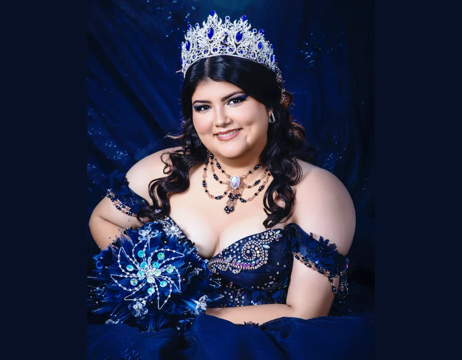 Quinceañera studio portrait in a navy blue off-the-shoulder gown with a jeweled bodice, layered necklace, and matching crystal tiara, holding a sparkling blue bouquet