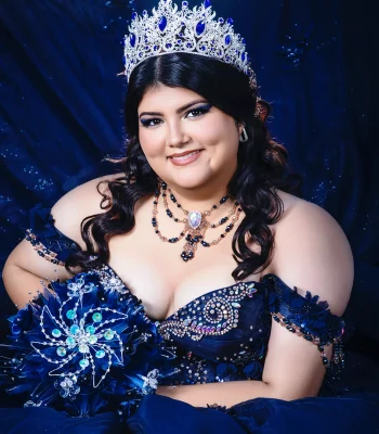 Quinceañera studio portrait in a navy blue off-the-shoulder gown with a jeweled bodice, layered necklace, and matching crystal tiara, holding a sparkling blue bouquet