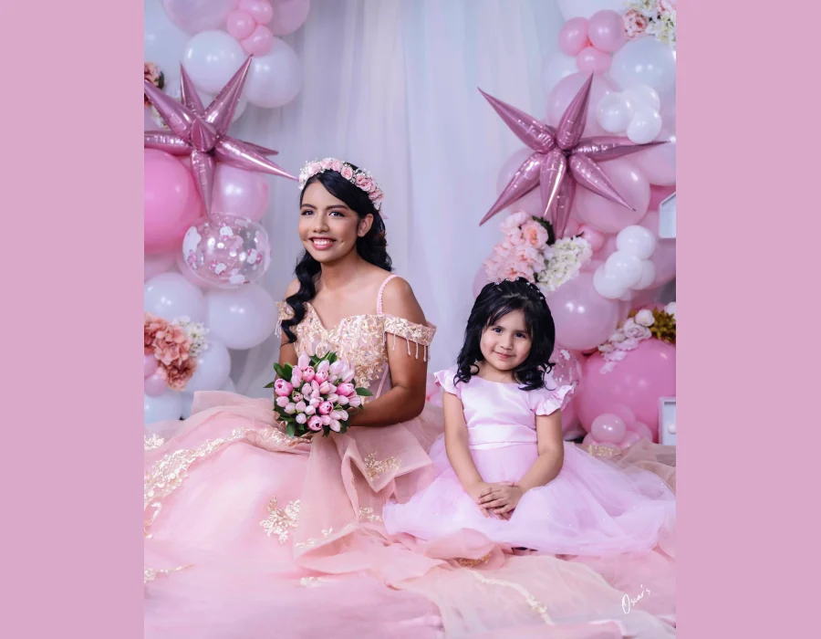 Quinceañera with her little sister in matching pink dresses, seated in front of a pink and white balloon arch with a tulle bouquet
