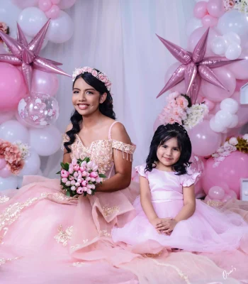 Quinceañera with her little sister in matching pink dresses, seated in front of a pink and white balloon arch with a tulle bouquet