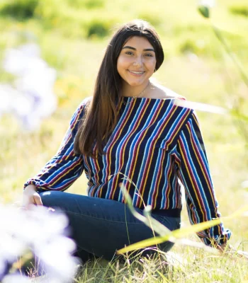 Quinceañera senior-style portrait in a striped sweater and jeans, sitting in a sunlit grassy field