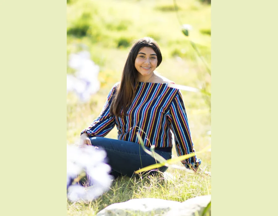 Quinceañera senior-style portrait in a striped sweater and jeans, sitting in a sunlit grassy field