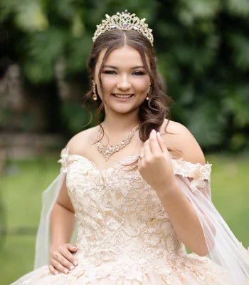 Quinceañera portrait in a champagne lace ball gown with a flowing veil and gold tiara, adjusting her hair in a garden setting
