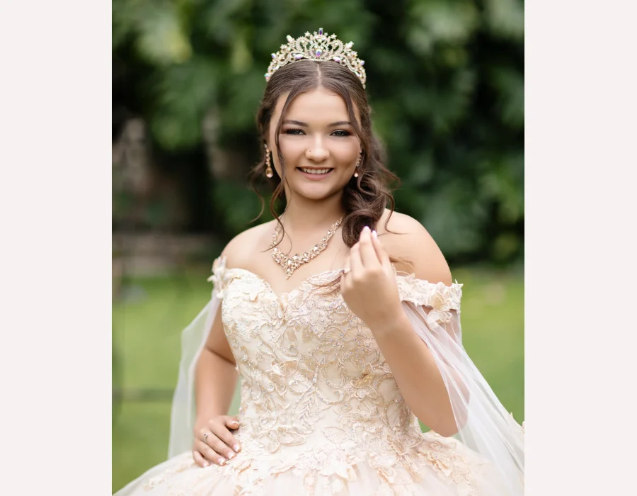 Quinceañera portrait in a champagne lace ball gown with a flowing veil and gold tiara, adjusting her hair in a garden setting