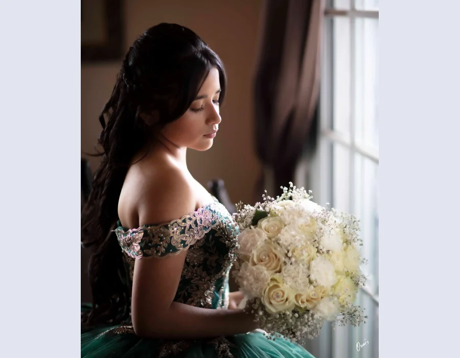 Quinceañera profile portrait in an emerald green off-the-shoulder gown with gold embroidery, holding a white rose bouquet by a window