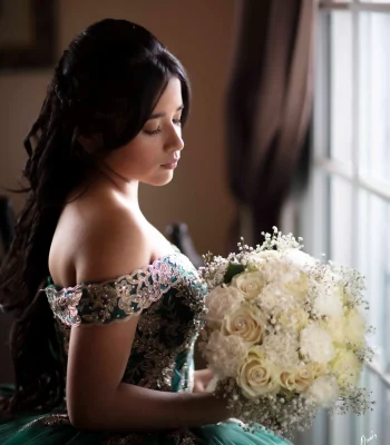 Quinceañera profile portrait in an emerald green off-the-shoulder gown with gold embroidery, holding a white rose bouquet by a window